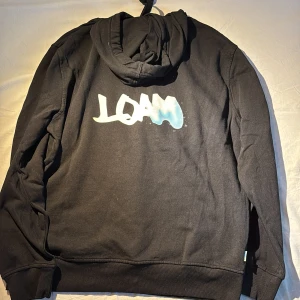 Svart hoodie med LOAM-tryck - Säljer en svart hoodie med ett coolt LOAM-tryck i vitt och blått på ryggen. Tröjan har en klassisk design med en stor ficka framtill och justerbar huva. Perfekt för en avslappnad stil.