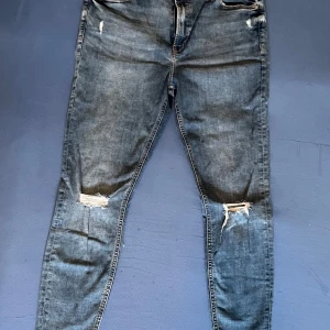 Blå jeans med slitningar - Snygga blå jeans med slitningar på knäna och låren. De har en klassisk femficksdesign och knappgylf. Perfekta för en avslappnad stil.
