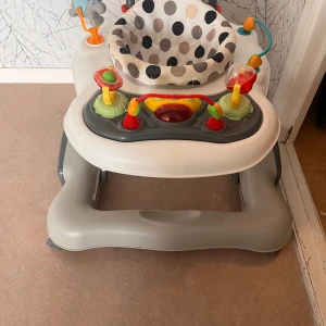 Gåstol med lekpanel - Säljer en praktisk gåstol i grått och vitt med en lekpanel. Sitsen har ett prickigt mönster i svart och beige. Lekpanelen har färgglada detaljer som röda, gula och gröna knappar samt leksaker. Perfekt för att underhålla och stödja barnets första steg.