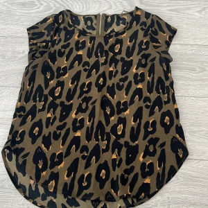 Leopardmönstrad blus med dragkedja - Snygg leopardmönstrad blus i jordnära färger med korta ärmar. Blusen har en praktisk dragkedja i ryggen för enkel på- och avtagning. Perfekt för att ge din outfit en vild touch!