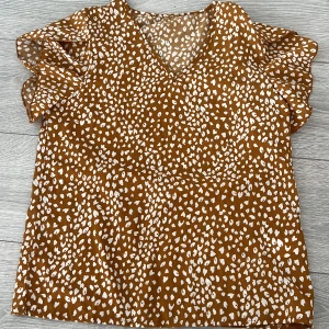 Brun och vit blus med volangärmar - Säljer en snygg brun blus med vita prickar och volangärmar. Blusen har en v-ringad hals och är perfekt för en trendig look.