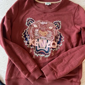 Röd sweatshirt från Kenzo med tigerbroderi - Säljer en snygg röd sweatshirt från Kenzo med ett stort broderat tigerhuvud och texten 'Kenzo Paris' på framsidan. Tröjan har långa ärmar och rund halsringning. Perfekt för en cool och avslappnad stil.