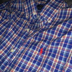 Rutig skjorta från Ralph Lauren - Säljer en snygg rutig skjorta från Ralph Lauren i blått, vitt och rött. Skjortan har en broderad logga på bröstet och knappar framtill. Perfekt för en stilren look!