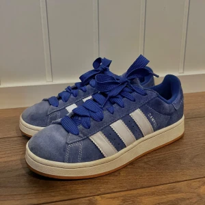 Adidas Campus 00s - Blå Adidas Campus i mocka med vita detaljer och klassiska tre ränder. Skorna har en rund tå och blå snörning. Bara använt ett fåtal gånger. 