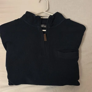 Mörkblå tröja från Dressmann - Säljer en stilren mörkblå tröja från Dressmann med halv dragkedja. Tröjan har en diskret bröstficka och är tillverkad i ett bekvämt material.