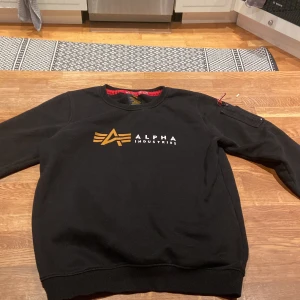 Svart sweatshirt från Alpha Industries - Säljer en svart sweatshirt från Alpha Industries med deras ikoniska logga i guld och vitt på bröstet. Tröjan har långa ärmar och en praktisk ficka med dragkedja på ärmen. Perfekt för en stilren och bekväm look.