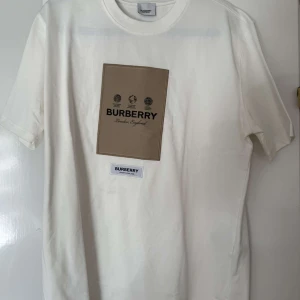 Vit t-shirt från Burberry - Säljer en stilren vit t-shirt från Burberry med ett stort beige tryck på framsidan. T-shirten är kortärmad och tillverkad i 100% bomull, vilket gör den bekväm och perfekt för vardagsbruk. Klassisk design med Burberrys logotyp och texten 'London, England'. Sommar Vibe