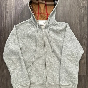 Grå hoodie från Burberry - Säljer en stilren grå hoodie från Burberry med dragkedja och klassiskt rutigt mönster i huvan. Tröjan har en broderad logga på bröstet och är perfekt för en avslappnad stil. Använd max 5 gånger.