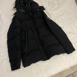 Vi kan diskutera priset privat - Säljer en svart dunjacka från Canada Goose med deras ikoniska märke på ärmen. Jackan har en dragkedja och knappar framtill samt en hög krage för extra värme. Perfekt för kalla vinterdagar.