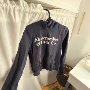 Mörkblå hoodie från Abercrombie & Fitch - Säljer en snygg mörkblå hoodie från Abercrombie & Fitch med vit logga på framsidan. Tröjan har långa ärmar och en klassisk huva. Perfekt för en avslappnad stil.
