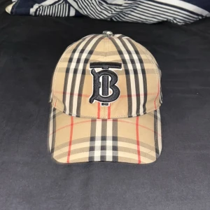 Burberry keps med broderad logga - Snygg Burberry keps i beige, svart och röd med en broderad logga framtill. Kepsen har justerbar rem baktill för perfekt passform. Inte använd. Pris kan diskuteras. står ej för frakt, kan mötas upp i GBG