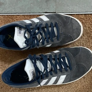 Adidas skor - Snygga Adidas sneakers i mörkblå/gråaktig mocka med de klassiska vita ränderna på sidan. Skorna har snörning och en rund tå, vilket ger en tidlös look. Perfekta för en avslappnad stil. Storlek 41 1/3 men känns större, passar 42 och antagligen t.o.m 43.