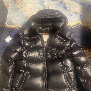 Svart pufferjacka från Moncler - Säljer en snygg svart pufferjacka från Moncler. Jackan har en quiltad design med dragkedja framtill och en huva för extra värme. Perfekt för kyliga dagar!