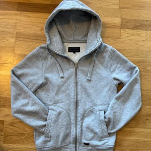 Grå hoodie från Dunderdon - Säljer en stilren grå hoodie från Dunderdon med dragkedja och justerbar huva. Perfekt för en avslappnad look. Den har praktiska fickor framtill och är tillverkad i mjukt material för extra komfort. Säljer pga för liten för mig.