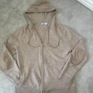 Beige kashmir hoodie från Rovoir - Säljer en stilren beige hoodie från Rovoir med dragkedja och huva. Tröjan har långa ärmar och snygga armbågslappar i matchande färg. Perfekt för en avslappnad stil.