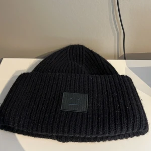 Svart ribbad mössa från Acne Studios - Snygg svart ribbad mössa från Acne Studios med ett stilrent ansiktsmärke framtill. Perfekt för att hålla värmen under kyliga dagar. Mössan har en klassisk design som passar till det mesta.