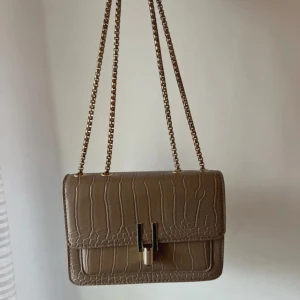 Beige axelväska med kedja - Snygg beige axelväska med krokodilpräglat mönster och guldfärgad kedja. Helt oanvänd!