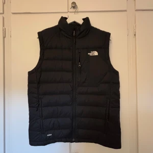 Svart dunväst från The North Face - Snygg svart dunväst från The North Face med dragkedja och logga på bröstet. Perfekt för kyliga dagar när du vill hålla värmen utan att kompromissa med stilen.