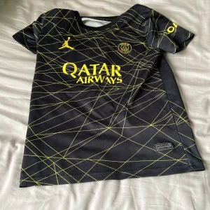 Tröja psg - Säljer en svart fotbollströja från Jordan med ett coolt gult mönster. Tröjan har korta ärmar och en snygg design med Qatar Airways-logga på framsidan. Perfekt för fotbollsfans som vill sticka ut på planen!