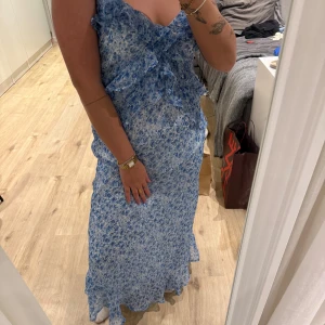 Blå blommig långklänning - Säljer en blå blommig långklänning från vero moda  med volangdetaljer. Klänningen har en omlott-design och är ärmlös, perfekt för sommarens festligheter. Den har en luftig passform och är gjord i ett lätt material. Har en defekt (se bild 4) därav priset, annars i väldigt fint skick. 