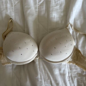 Vit Pusch up bh med spets och strass från H&M - Säljer en elegant beige bh från H&M med vacker spetsdetalj och strass på kuporna. Bh:n har justerbara axelband och är tillverkad i en mjuk polyesterblandning. Perfekt för att känna sig extra fin! Väl använd 75A
