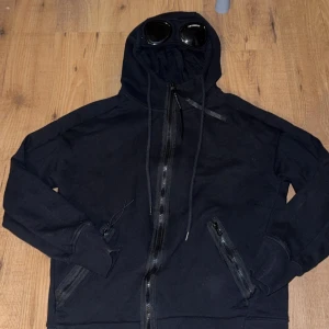 Svart hoodie med glasögon från C.P. Company - ‼️använd 1 gång‼️Säljer en cool svart hoodie från C.P. Company med inbyggda glasögon i huvan. Den har dragkedja framtill och snörning vid huvan. Perfekt för en unik och stilren look. 