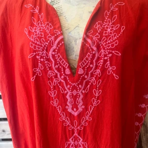 Röd kaftan med broderi - Säljer en vacker röd kaftan med rosa broderade detaljer och tofsar. Den har en v-ringad hals och är dekorerad med blommönster längs ärmarna och framsidan. Perfekt för en bohemisk look. Använd någon enstaka gång och tvättas den gång, super fint skick. Nypris 799:-