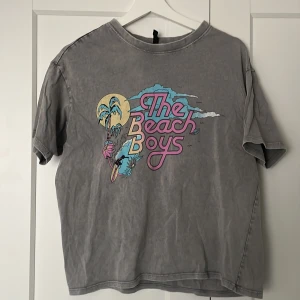 Grå t-shirt med The Beach Boys-tryck från H&M - Säljer en grå t-shirt från H&M med ett färgglatt The Beach Boys-tryck på framsidan. T-shirten har en avslappnad passform och korta ärmar, perfekt för en casual look. Materialet är mjukt och bekvämt.