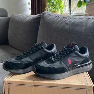 Svarta sneakers från Polo Ralph Lauren - Snygga svarta sneakers från Polo Ralph Lauren med röd logga på sidan och tungan. Skorna har en kombination av skinn och mocka med snörning framtill. Perfekta för en stilren look. Knappt använda använt ca 3 gånger, riktigt bra skick och superbekväma. Säljer på grund av att jag inte använder de. Hör av dig vid frågor eller funderingar!