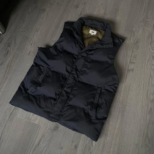 Mörkblå dunväst - Säljer en stilren mörkblå dunväst frånmärket WE köpt på Uniqlo. Västen har dragkedja och knappar framtill samt två praktiska fickor. Perfekt nu inför vår och sommar. Västen är i mycket fint skick förutom ett hår i armhålan på ena sidan ( se på sista bilden ). Går säkert att lösa om man känner någon som kan sy. Overall en mycket fin och fräsch väst. Köpt för 800kr. Storlek S men passar minst lika bra på M