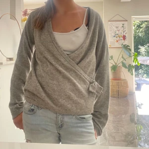 Grå omlottkofta  - Mysig grå omlottkofta med v-ringad hals och en söt rosettdetalj på sidan. Koftan har långa ärmar och ribbade muddar vid nederkant och ärmslut. Perfekt att slänga på över ett linne för en chill och stilren look.