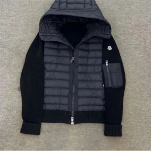 Moncler cardigan - Snygg svart pufferjacka från Moncler med stickade ärmar och huva. Jackan har quiltad front, dragkedja och en cool ficka med zip på ärmen. Perfekt för dig som gillar streetstyle och vill ha något som sticker ut. Moncler-logga på axeln för extra vibe.