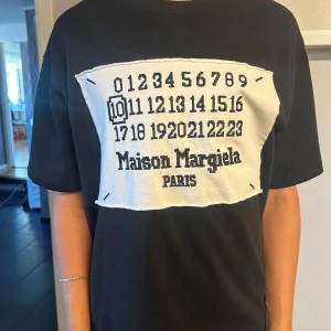 Maison Margiela t-shirt  - Svart t-shirt från Maison Margiela med ikoniskt vitt patch och siffror samt logga på bröstet. Rund hals och korta ärmar. Tillverkad i mjuk bomull, klassisk passform och cool kontrast mellan svart och vitt. Bra skick på plagget då den knappt är använd. Storlek S.