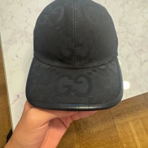 Svart Gucci keps med GG-mönster - Snygg svart keps från Gucci med det klassiska GG-mönstret ton-i-ton över hela kepsen. Kepsen har böjd skärm och justerbar rem baktill. Materialet är slitstark bomull med detaljer i syntet. Perfekt för dig som vill ha en lyxig streetstyle-look.