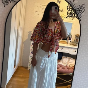 Blommig omlott blus med knyt - Säljer en färgglad blommig blus med korta ärmar och djup v-ringning. Toppen har omlott-design och knyts framtill, vilket ger en croppad och luftig look. Mönstret är i rött, gult, rosa och grönt på vit botten. Perfekt för sommarens alla häng!