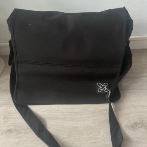 Svart skötarväska med axelrem - Svart skötarväska med justerbar axelrem och flera fack, både med dragkedja och tryckknapp. Väskan har meshficka på sidan och ett stilrent emblem framtill. Insidan har organiseringsfack och elastiska band för småsaker. 