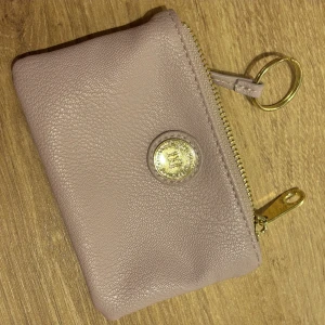 Beige skinnplånbok med gulddetaljer - Snygg beige plånbok i äkta skinn med guldfärgad dragkedja och logga framtill. Perfekt storlek för kort och mynt, med en praktisk nyckelring i guld. Stilren och enkel design som passar i fickan eller väskan.