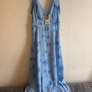 Blommig blå långklänning Bik Bok - Säljer en ljusblå långklänning från Bik Bok med v-ringad hals och breda axelband. Klänningen har ett fint blommigt mönster i olika blå nyanser och är gjord i ett mjukt, flowy material som känns som polyester. Perfekt för sommarens alla tillfällen!