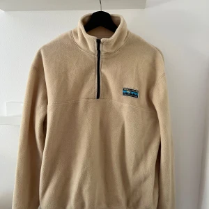 Beige fleece-tröja från Junkyard - Säljer en beige fleece-tröja från Junkyard i storlek XS. Tröjan har half zip framtill, hög krage och ett litet Urban Tears-märke på bröstet. Tillverkad i mjuk polyester som är perfekt för kyliga dagar. Långärmad och loose fit för skön och avslappnad stil.
