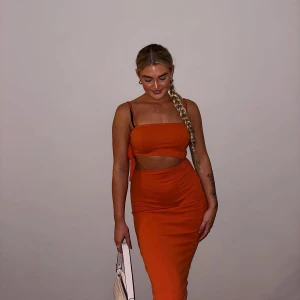 Orange omlott långklänning med cutout - Snygg orange långklänning med smala axelband och cool cutout-detalj i midjan. Klänningen har omlott-design på sidan och sitter tight mot kroppen för en riktigt trendig look. Perfekt för dig som gillar att sticka ut med färg och form.