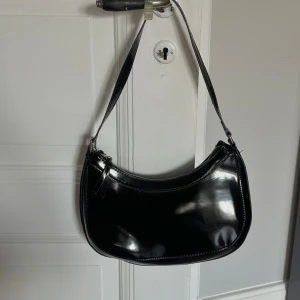 Svart axelväska i blankt skinn - Snygg svart axelväska i blankt skinn med rundad form och silverfärgad dragkedja upptill. Väskan har ett stilrent och minimalistiskt utseende som passar perfekt till din outfit. Axelremmen är bred och bekväm, och väskan har en modern vibe.