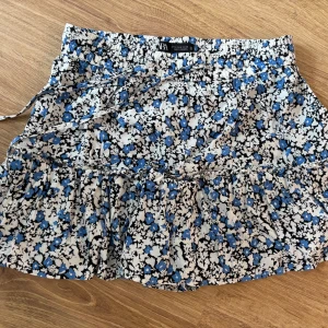 Blommig kortkjol från Zara - Supersöt kortkjol från Zara med blommigt mönster i blått, svart och vitt. Kjolen har volang och en smockad midja med knytband. Perfekt för dig som gillar färg och mönster! Materialet känns lätt och luftigt.