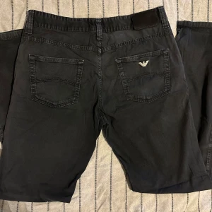 Svarta chinos från Armani Jeans - Snygga svarta chinos från Armani Jeans med klassisk femficksdesign och silverfärgad logga på bakfickan. Byxorna har raka ben, normal passform och är tillverkade i mjuk bomull. Perfekta för dig som gillar stilrena och exklusiva detaljer.