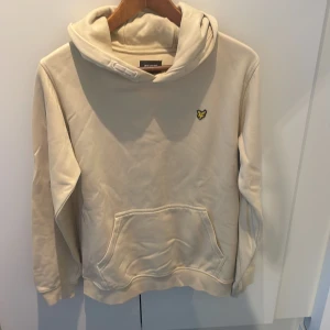 Beige hoodie från Lyle & Scott - Säljer en beige hoodie från Lyle & Scott med klassisk gul fågel-logga på bröstet. Tröjan har huva, magficka och långa ärmar. Materialet är mjuk bomull blandat med polyester, perfekt för chill dagar. Enkel och stilren design som funkar till det mesta.