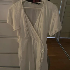 Vit omlottklänning från Vero Moda - Snygg vit kortklänning från Vero Moda perfekt på student eller Konfa klänningen är i omlottmodell med knytband i midjan. Klänningen har korta ärmar och är tillverkad i mjuk bomull, vilket gör den superbekväm. 