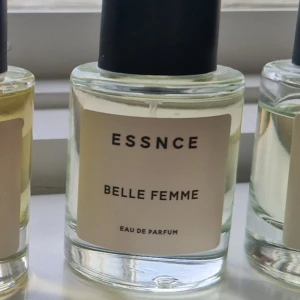 Essnce Belle Femme Eau de Parfum, 50 ml - Essnce Belle Femme Eau de Parfum i en stilren, rund glasflaska med svart lock. Parfymen har en transparent, ljusgul ton och minimalistisk etikett med svart text. Perfekt för dig som gillar enkel och modern design. Testsprutad ca 2-3 sprut.