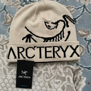 Beige Arc'teryx mössa med logga - Snygg beige mössa från Arc'teryx med svart fossil- och logotypdesign. Mössan har uppvik och är stickad i mjukt material som håller dig varm. Perfekt för dig som gillar outdoor och vill ha en clean look med tydlig branding.