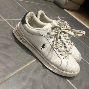 Vita sneakers från Polo Ralph Lauren - Snygga vita sneakers från Polo Ralph Lauren med marinblå detaljer och logga på sidan. Skorna har rund tå, platt sula och snörning med Polo-tryck på skosnörena. Tillverkade i skinn med mjuk textil på insidan. Perfekt för dig som gillar klassisk och sportig stil. Det gröna är färg men de går säkert bort! 