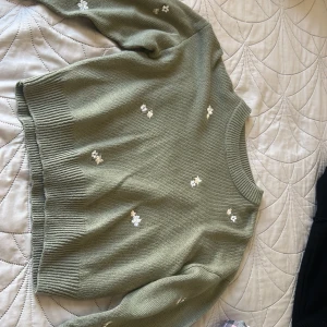 Olivgrön stickad tröja med blommor - Olivgrön stickad tröja från H&M Divided i storlek S. Tröjan har broderade vita blommor spridda över hela plagget och ribbade muddar vid ärmslut och nederkant. Rund hals och långärmad modell