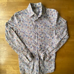 Blommig skjorta från Bläck, tailored fit - Säljer en snygg blommig skjorta från Bläck i tailored fit. Skjortan har ett färgstarkt mönster med blå, röda och gröna blommor på vit botten, klassisk krage och långa ärmar. Perfekt för dig som gillar att sticka ut med stilrena detaljer och knappar framtill.